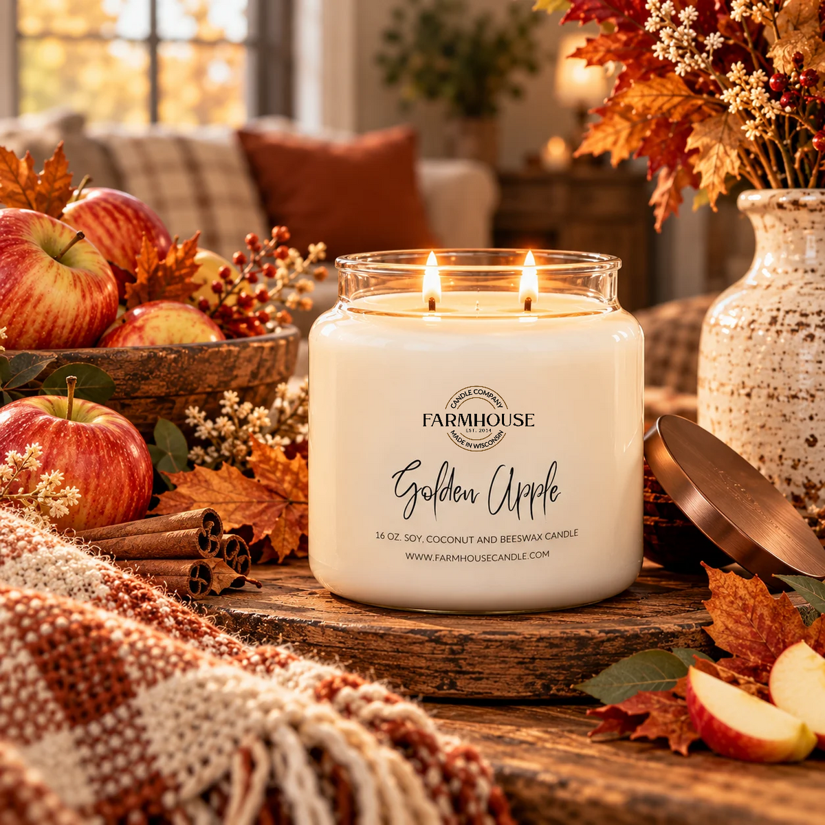 A lit Farmhouse Candle Company 'Golden Apple' scented candle in a clear glass jar sits on a rustic wooden table surrounded by fall-themed props including red apples, cinnamon sticks, dried leaves, and a cozy throw blanket.