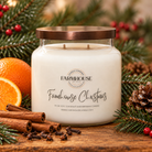 A Farmhouse Candle Company brand candle labeled 'Farmhouse Christmas' made with soy, coconut and beeswax in a glass jar with bronze lid.  Pine cones, boughs, cinnamon, cloves and oranges on wood.